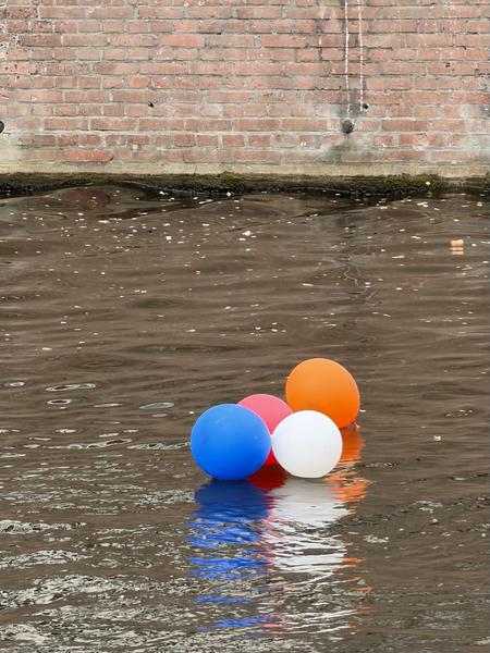 Four colorful balloons in blue, red, white, and orange float gently on the rippling brown water of a canal